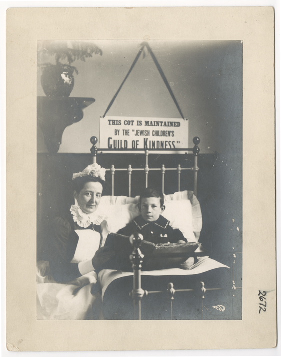 Black and white photo of a nurse sat with a child in a cot. They're looking to camera but have a big book that they are reading.
