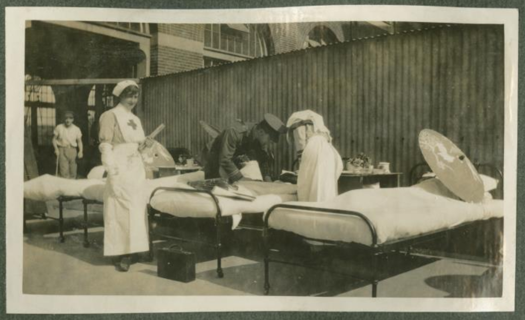 Black & white photo of nurses attending beds & soldiers in the open air. Metal beds are by a wooden fence in front of a brick building