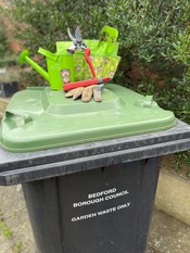 A green lidded bin with gardening tools on the lid. A green bush in the background.