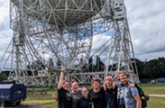 A group of staff stand together in front of the Lovell Telescope at blue dot. They are all looking towards the camera and smiling. 