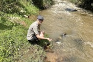A picture of PhD student Sophia crouching by a river, taking samples for her research. She wears a shirt, shorts and a hat. 
