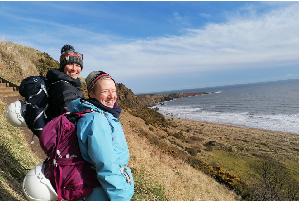Two female geologists stand by the sea. They are wearing technical backpacks with helmets attached
