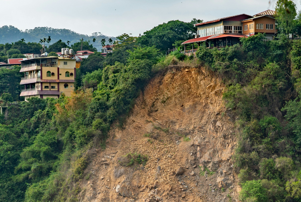 Landslide aftermath, showing the slip from the top of a cliff. Houses perches atop the cliff. 