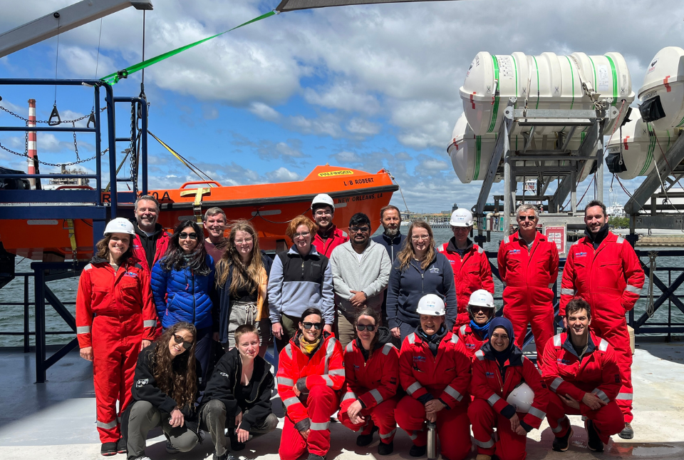 The IODP team stand together on the ship's deck. Some are wearing casual clothes, others are in red boilersuits and white hard hats.