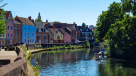 The rvier Wensum in Norwich, Norfolk, England. You can see people on the water in canoes, and the houses by the river are multicoloured