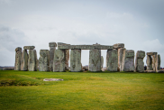 An image of Stonehenge on a cloudy day. The grass below is green, and a bit scorched in places. 
