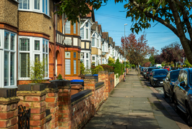 A row of houses in London, with a pavement in the middle of the image and cars parked on a road to the right hand side.  