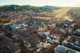 An aerial image of Bologna during the day. Shafts of light can be seen over the buildings rolling hills are in the background