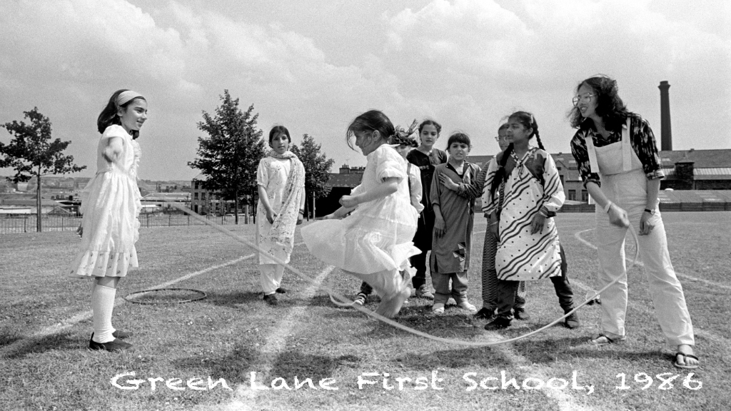A group of young girls outside in a playground, skipping.  It is subtitled 'Green Lane First School 1986'
