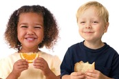 Photograph of a girl eating a slice of orange and a boy with a slice of wholemeal bread