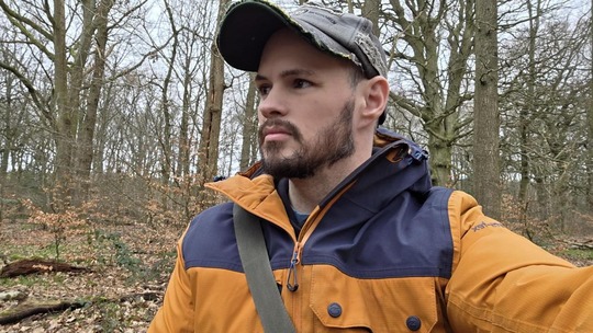 man in a cap and mustard yellow coat with blue panels on the shoulders in the woods