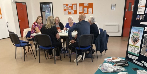 customers sat around a table chatting in a community hall- Information table to the right in the foreground