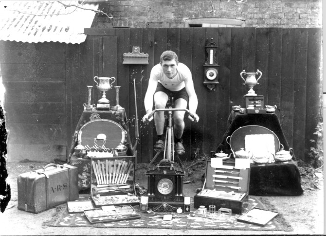 Historical photograph of sportsman cyclist, studio photograph of cyclist on bicycle facing camera, surrounded by trophies