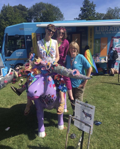 library staff at Cambridge Pride 2022. Two members of staff are sitting on the Cambridge Pride cow. There's a mobile library in the background.