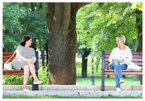 Two women sitting on separate benches in a park smiling at each other 
