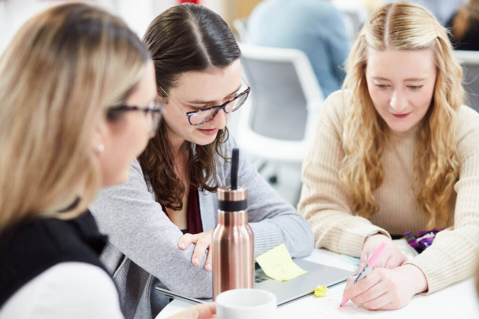 A woman writes with a pen in a group setting with two colleagues 