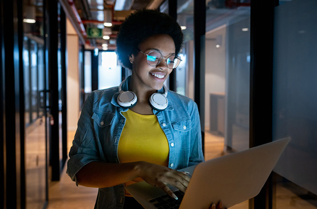 A woman wearing glasses, with headphones around her neck is smiling and holding a laptop  
