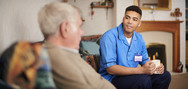 Male carer in uniform sits on sofa with mug chatting to elderly man in foreground with head turned away from camera