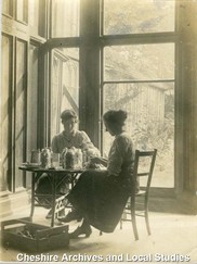 Very old black and white photograph shows two ladies in Edwardian dress taking tea at a table near a large window