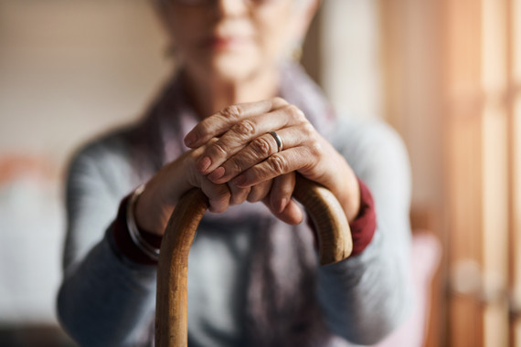 A pair of older lady's hands sit on top of a curved walking stick in the foreground. The background is a blurry outline of a lady. 