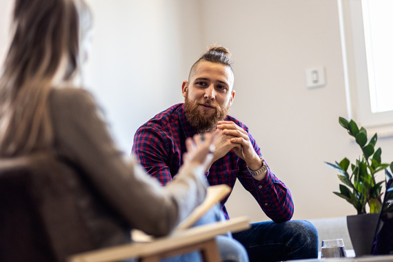 Man with long hair in a bun sits in chair opposite woman