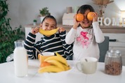 Two children at a kitchen counter holding fruit up in front of their faces