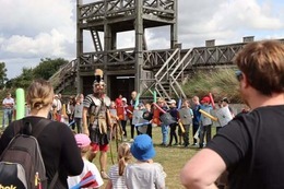 Lunt Roman Fort in the background with a Roman in front of it and a crowd surrounding him