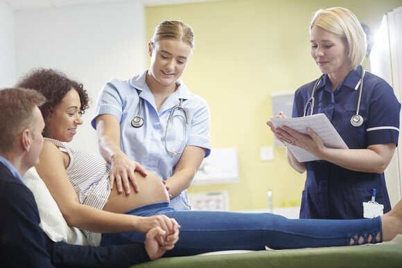 Photograph of two healthcare professionals checking and observing a pregnant mother who is lying on a hospital bed. Their partner comforts them.