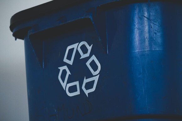 Close-up photograph of a blue recycling bin, which shows three white line arrows arranged in a triangle to represent 'recycle' on the side