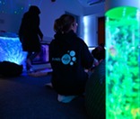 Person sat on floor facing way wears a hoodie with 'Cumberland Family Hubs' next to bubble tube in dark sensory room