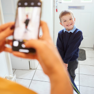 Boy stands in front of door in school uniform smiling, out of focus phone takes a picture