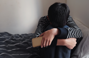 young child sitting on bed hugging knees with head down