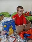 Baby sits in box next to the book Whatever Next, the cover of the book shows a bear in a cardboard box