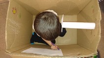 Boy sat inside a cardboard box using a felt-tip pen to write on the inside of the box