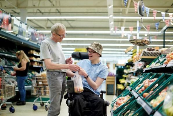 A man supporting a disabled person in a wheelchair with his shopping.