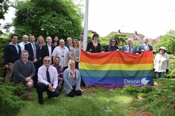 Nineteen people standing by the flagpole. Several are holding a large rainbow flag that includes Devon County Council's logo