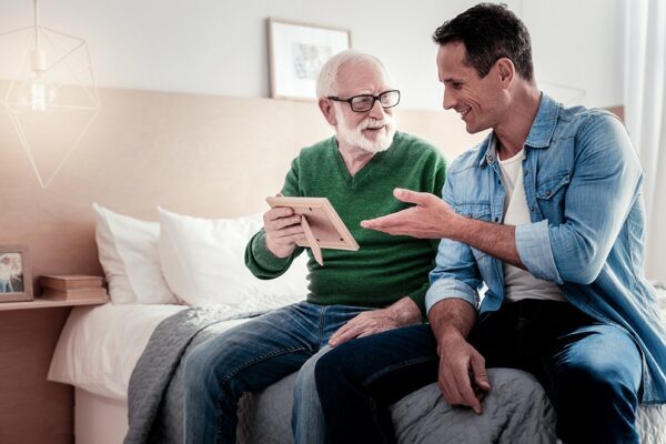 An elderly gentleman sitting by his son on a bed. They are smiling and looking at a framed photograph.