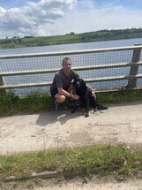 Darren, a middle age man with dark hair, kneeling on a bridge looking at the camera. To his left sits Ruth a black Labrador Retriever Guide Dog.