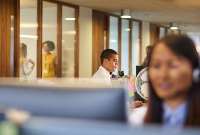 Blurred image of a woman sat at her computer in an office setting. People in the office are working behind her