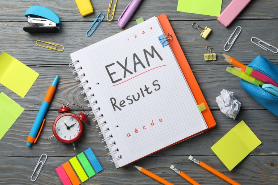 The words 'exam results' on a colourful piece of card, on a desk on which there is a range of items, including pens, stapler