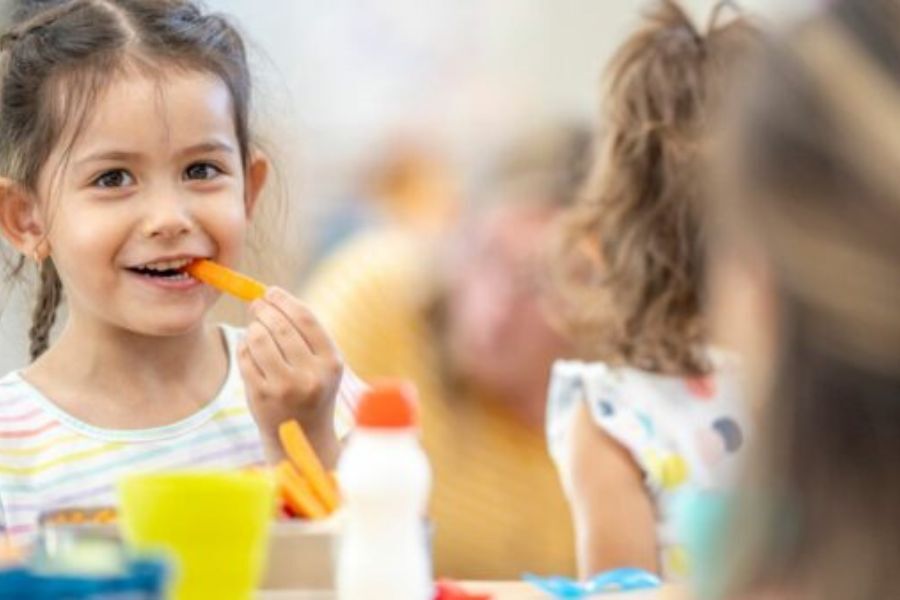 A young girl, smiling, and eating food at a table with other children