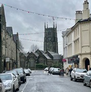 Image of a residential street with cars parked either side and a church with a tower at the end.