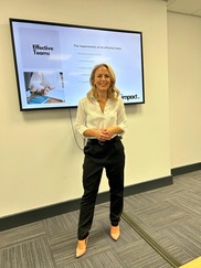 A white woman in black trousers and a white shirt with heeled shoes in front of a screen which says effective teams.