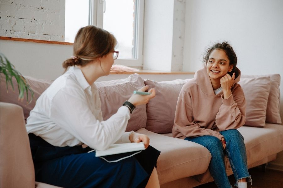 A teenager sat on a settee talking to an adult professional, smiling.