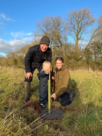 A Free Tree Scheme applicant with her family, an older male and toddler. Stood proudly around the tree they planted on their land.