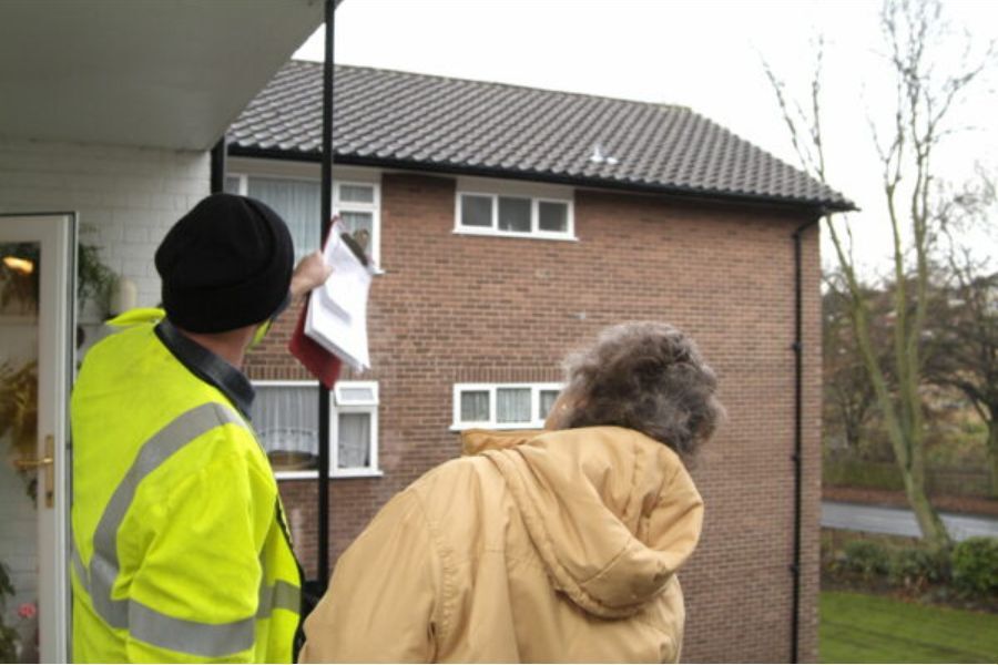 A man in high-vis jacket talking to an elderly woman on her doorstep, pointing up at her roof