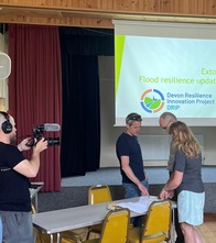 Three people stood around a table, pointing at some paper and discussing, whilst being filmed by someone with a camera