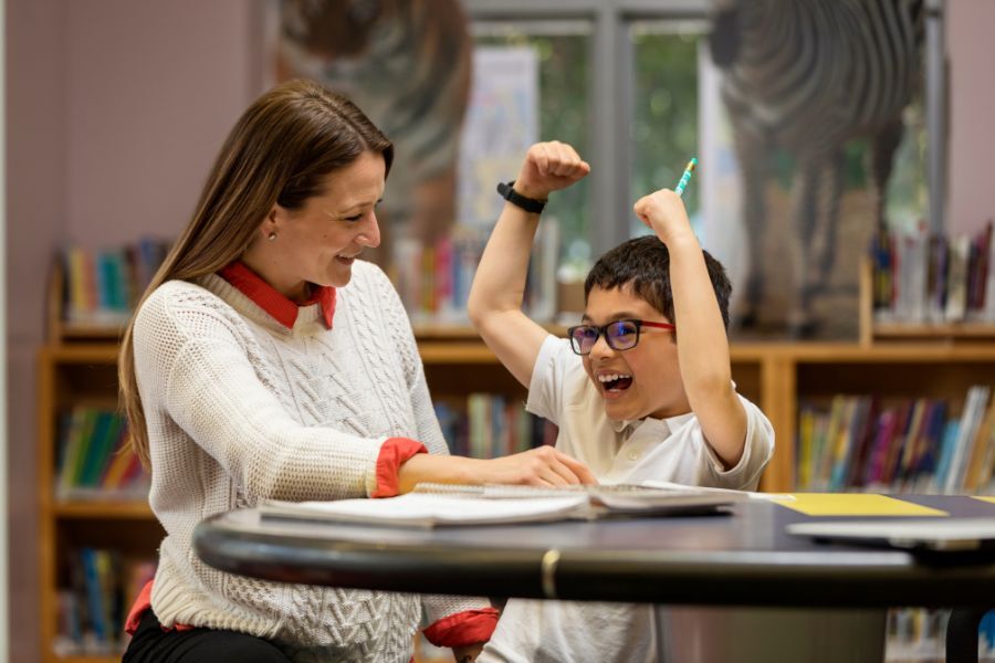 An adult sitting with a pupil in a classroom at a table, helping the pupil with his reading.