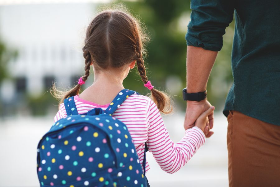 A parent holding his young daughter's hand as they go to school. The girl is carrying a bag on her back.