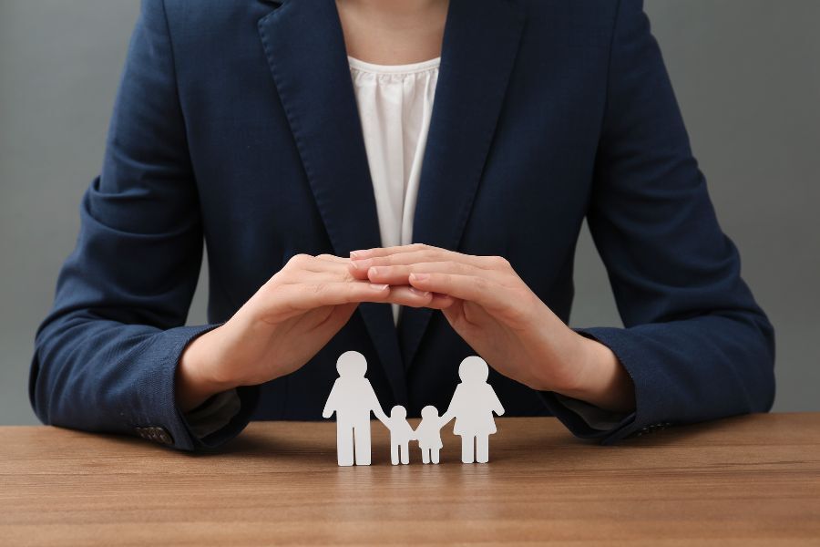 Image shows a professional person sat at a table holding her hands in a protective way above a paper cut-out representing a family